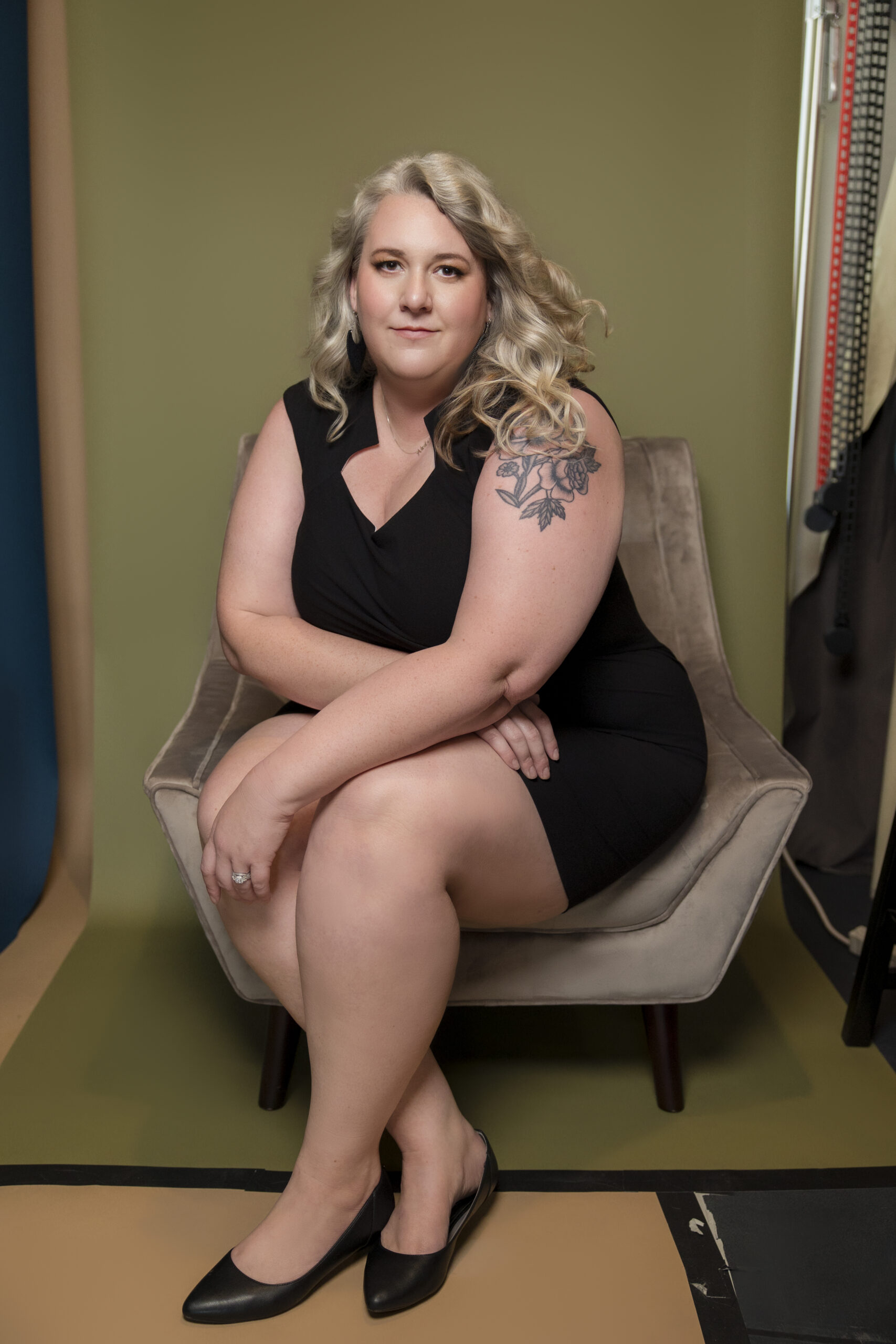 Nicole Howser sitting in a low-back chair against an olive backdrop, fan blowing her curls, posture strong and gaze calm.